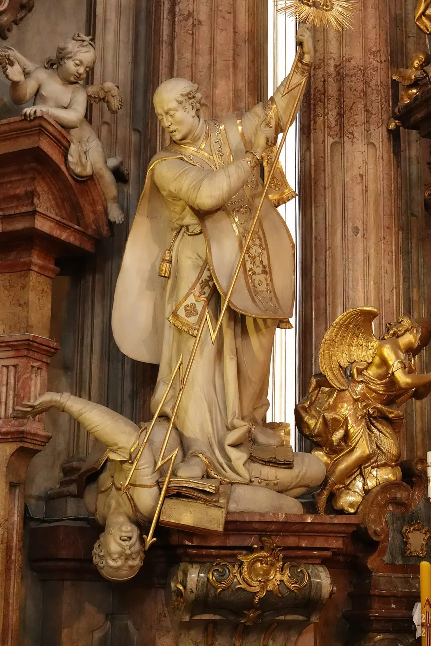 Statue of St. Ignatius of Loyola raising the sunburst of the Holy Name and driving his staff down onto a heretic who falls headlong with his book